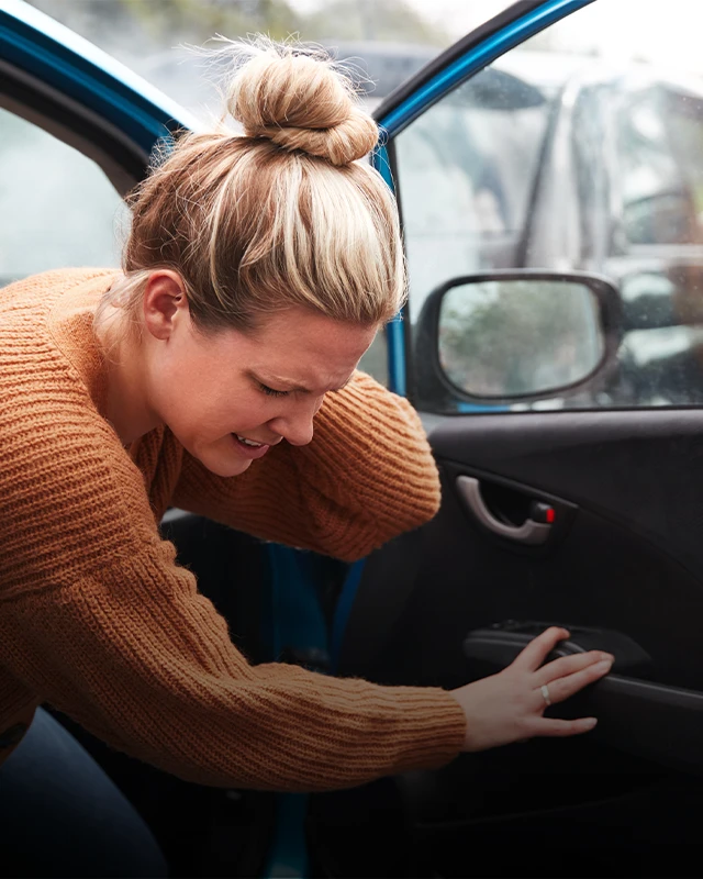 Person wearing a brown knitted sweater leaning into the open door of a blue car, with one hand on the car door handle and the other resting inside the vehicle