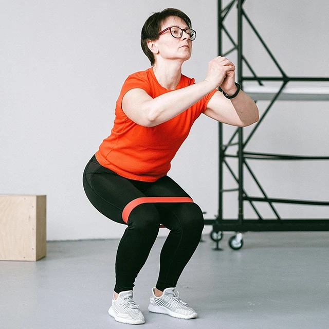 Person performing a squat exercise indoors using a red resistance band around the thighs, wearing a bright orange shirt and black leggings