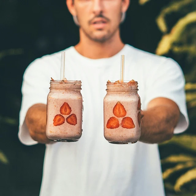Person holding two glass jars filled with strawberry smoothies, each decorated with sliced strawberries on the outside and topped with a straw