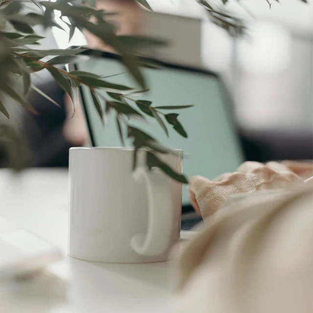 Close-up of a white coffee mug on a desk with green plant leaves in the foreground and a laptop in the background