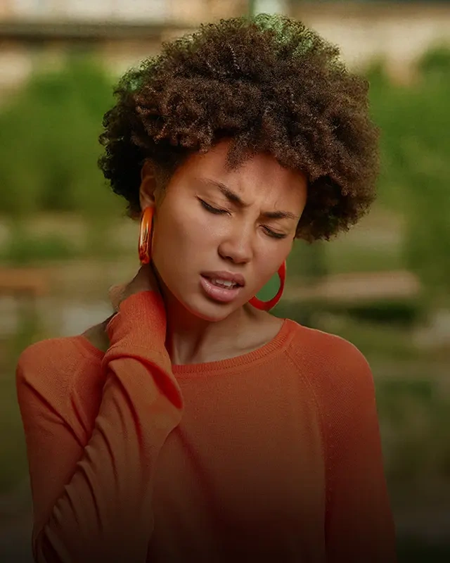 Person wearing an orange long-sleeve sweater standing outdoors, with one hand resting on the neck and curly hair visible against a green background