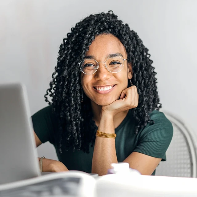 Woman sitting at a desk with a laptop, wearing a dark green short-sleeve top and a bracelet, resting their chin on one hand