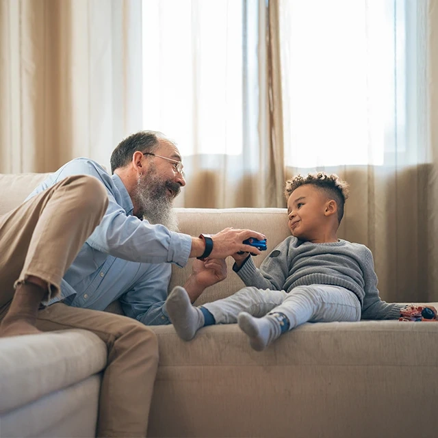 Adult and child sitting on a beige sofa in a living room, interacting with a video game controller