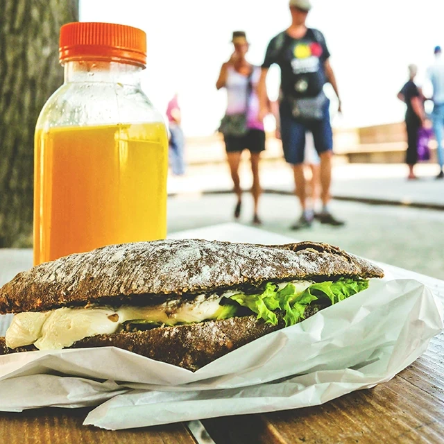 Close-up of a rustic sandwich with lettuce and cheese on a wooden table, next to a bottle of orange juice, with people walking in the background