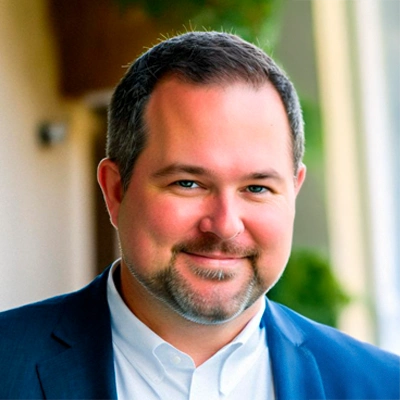 Darin Schultz wearing a dark suit jacket over a white dress shirt, standing in an outdoor setting with blurred greenery and architectural elements in the background
